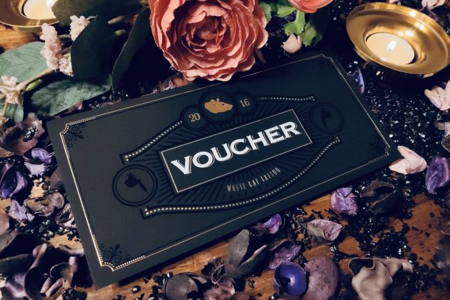 Czarny voucher White Rat Tattoo pośród kwiatów, świec i płatków – elegancki prezent na tatuaż w Warszawie 3